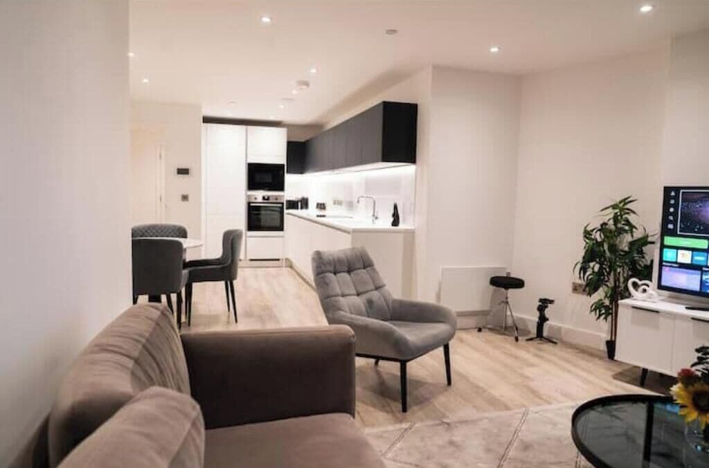 A modern living room with a sofa, armchair, and a small table with a vase. A kitchen is visible in the background with a dining area.