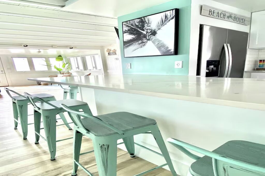 Een keuken met een groot eiland, barkrukken, een koelkast, en een bord aan de muur met de tekst 'BEACH HOUSE'.