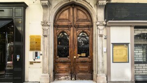 Une porte en bois à deux battants, ornée d’une plaque, une porte noire à gauche et un mur de briques à droite.