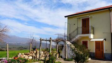 Ein zweistöckiges Gebäude mit einem Balkon, einem Garten mit Topfpflanzen und einer Berglandschaft im Hintergrund.