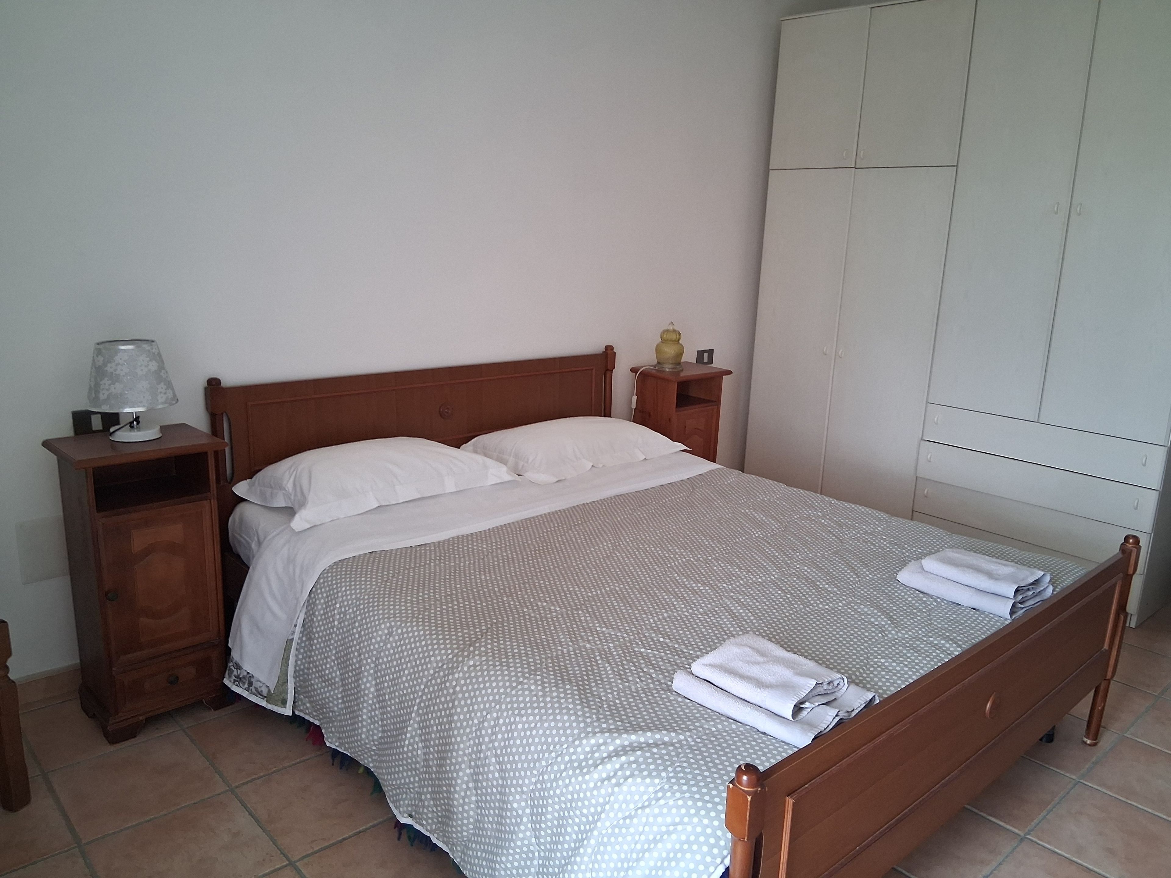 A single bed with a wooden frame, a nightstand with a lamp, and a wardrobe against a plain wall.