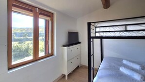 Une chambre avec un lit superposé, une fenêtre donnant sur un paysage verdoyant, une commode blanche avec un téléviseur à écran plat et une poutre en bois au plafond.