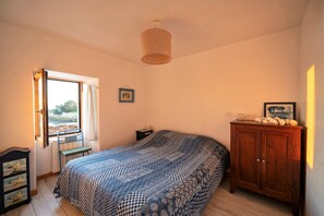 Ein Schlafzimmer mit einem Bett, einem Holzschrank, einem Sessel, einem kleinen Tisch, einem Bild an der Wand und einem Fenster mit Vorhängen.
