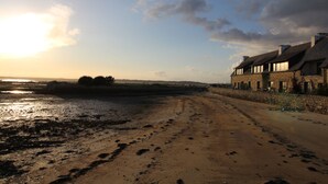 A coastal road leading to a stone building with a sloped roof, situated beside a sandy beach with footprints. - One of a kind! Ground-floor apartment with a terrace overlooking the sea (Carnac)