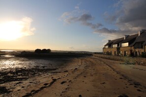 Eine Küstenstraße führt zu einem Steinhaus mit geneigtem Dach, das neben einem Sandstrand mit Fußabdrücken liegt.
