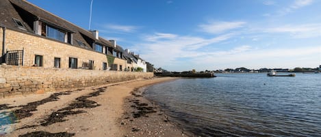 Un bâtiment situé sur la côte, doté de murs en pierre et de nombreuses fenêtres, et jouxtant une plage de sable où l’on trouve des algues et des débris.