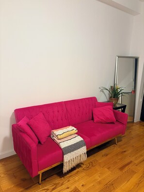 A red sofa with a striped throw blanket and pillows, positioned against a white wall with a wooden floor. - Bright apartment in LiC/Astoria fully equipped with balcony and skyline views (Astoria)