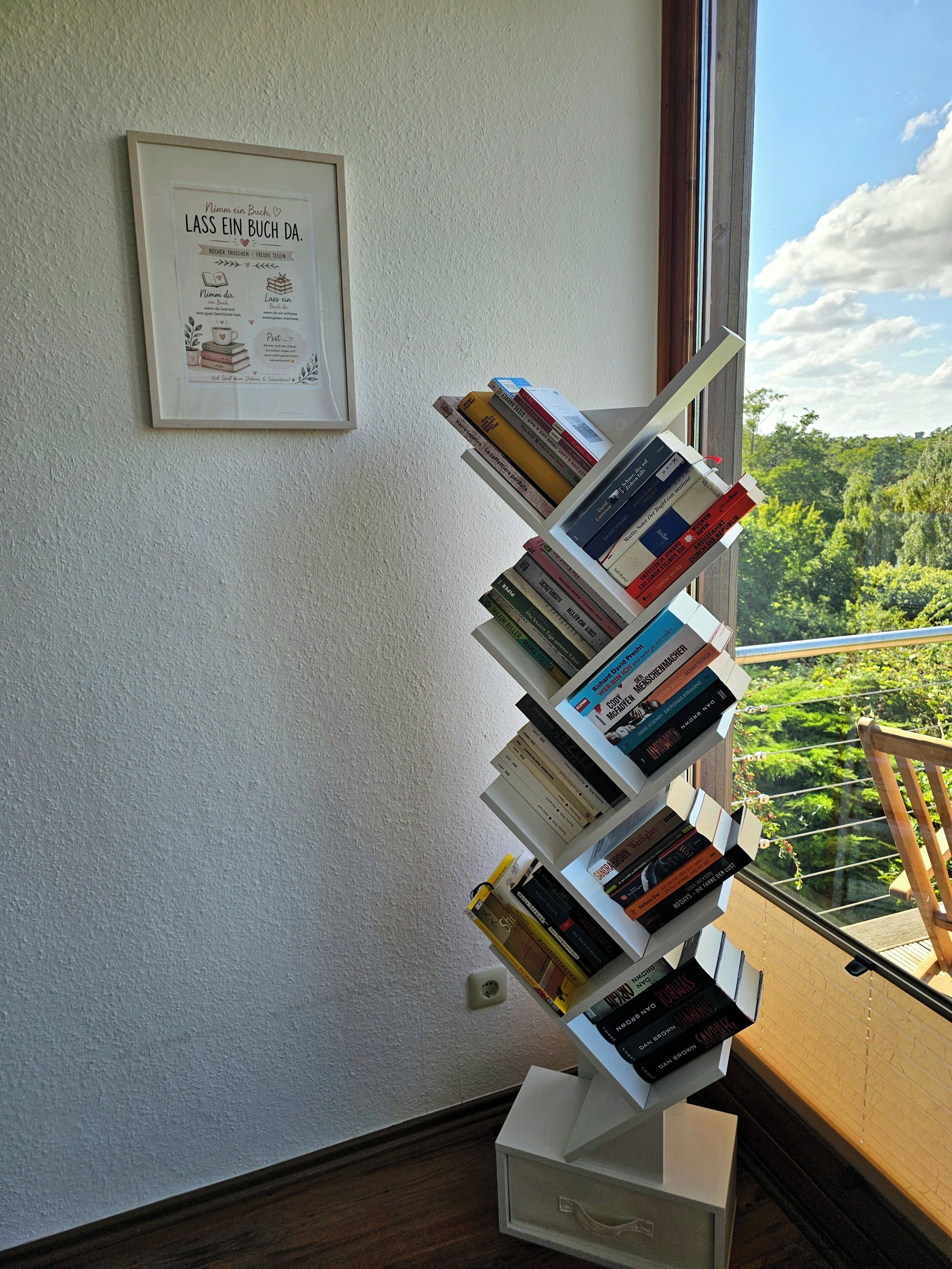 Uma estante com livros, um quadro na parede e vista para o verde lá fora.