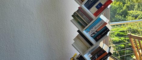 Uma estante com livros, um quadro na parede e vista para o verde lá fora.