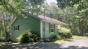 A green single-story house with a gravel driveway, surrounded by trees and shrubs. - Two-Bedroom One-Bathroom Vacation Home - Pet Friendly! (Hunts Point)