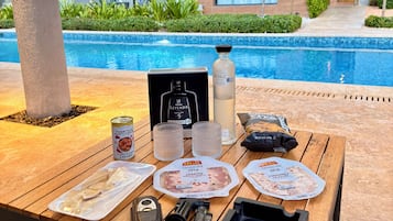A poolside table with food items, a bottle, and a box, set against a backdrop of a modern building and a swimming pool.