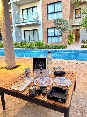 A poolside table with food items, a bottle, and a box, set against a backdrop of a modern building and a swimming pool.