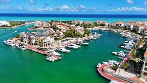 A marina with multiple yachts docked, surrounded by buildings with red-roofed structures, and a clear view of the ocean.