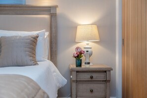 A neatly made bed with a headboard, a bedside lamp, a small wooden nightstand with drawers, and a vase with flowers. - The old School House Sleeps 16 (Barnoldswick)