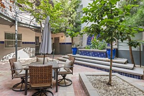 Eine Terrasse im Freien mit Esstisch, Stühlen und Sonnenschirm. Treppen führen zu einer Sitzgelegenheit mit blauen Fliesen und Pflanzkübeln.