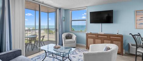 A living room with a glass table, a sofa, a chair, a TV, and a view of the ocean.