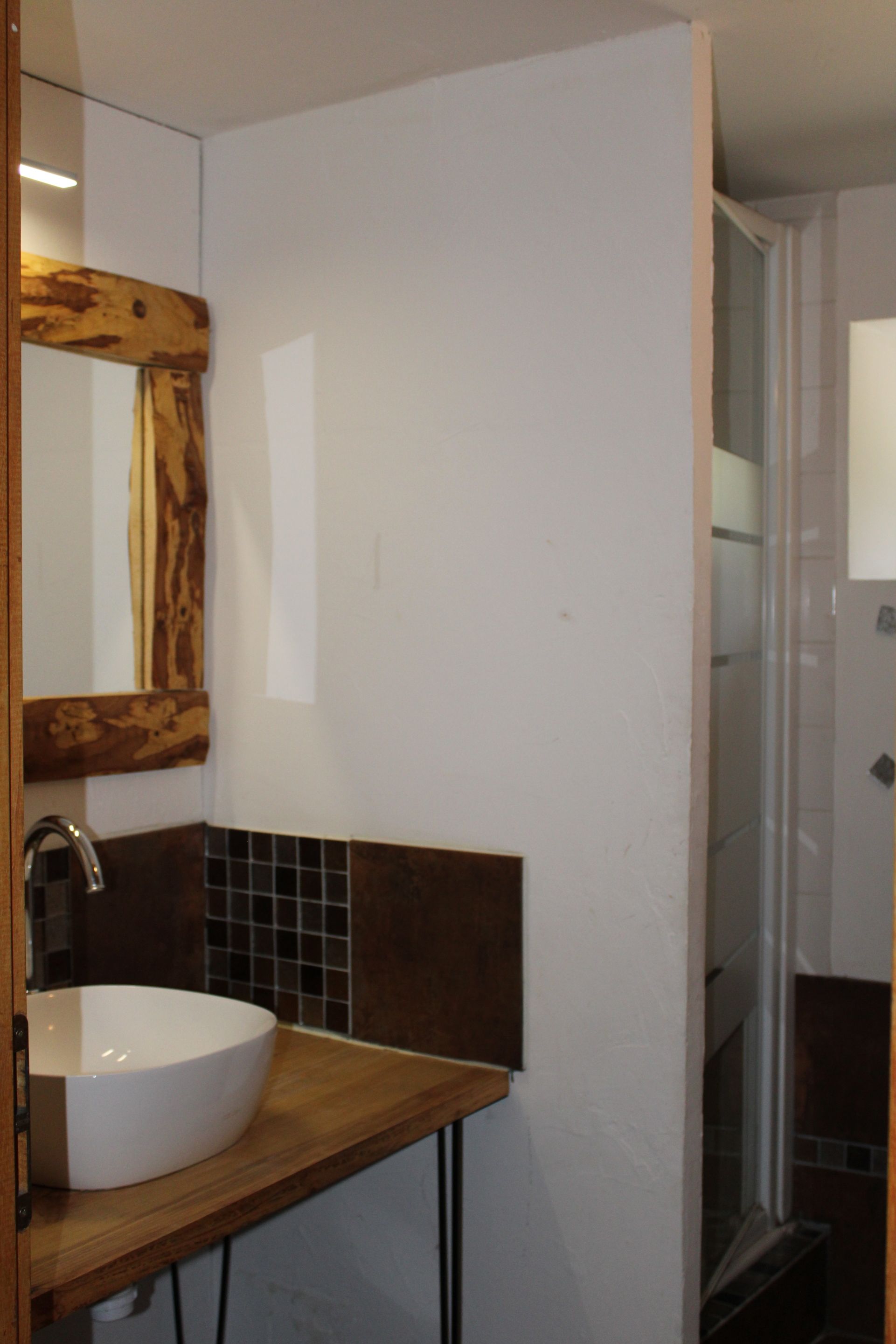 Ein Badezimmer mit einem Holzbadezimmerhocker, einem weißen Waschbecken, einem Spiegel mit Holzrahmen und einem gefliesten Duschbereich.