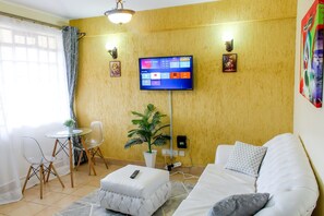 A living room with a sofa, a small table, a chair, a potted plant, and a TV mounted on the wall. - Cijana Gardens - School lane 1 br Westlands (Nairobi)
