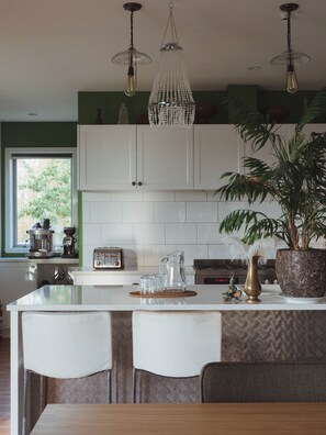 Een keuken met een centraal eiland, witte kasten, een hanglamp en een potplant.