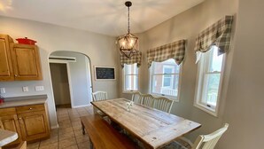 A dining area with a wooden table, benches, and white chairs. There is a chalkboard on the wall and a window with plaid curtains.