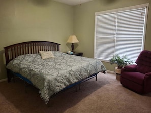 A bedroom with a bed, a nightstand, a lamp, a window with blinds, a chair, and a potted plant.