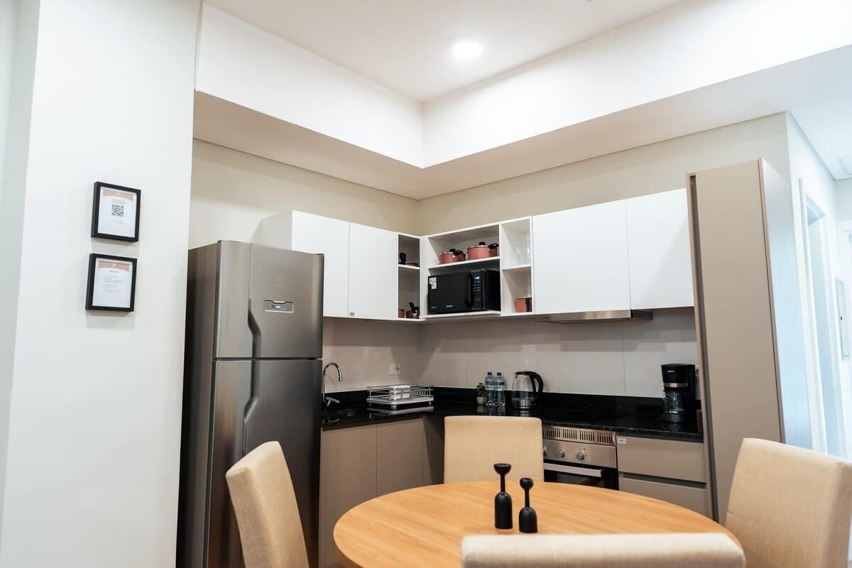 A compact kitchen with white cabinets, a stainless steel refrigerator, a round wooden table, and two chairs.