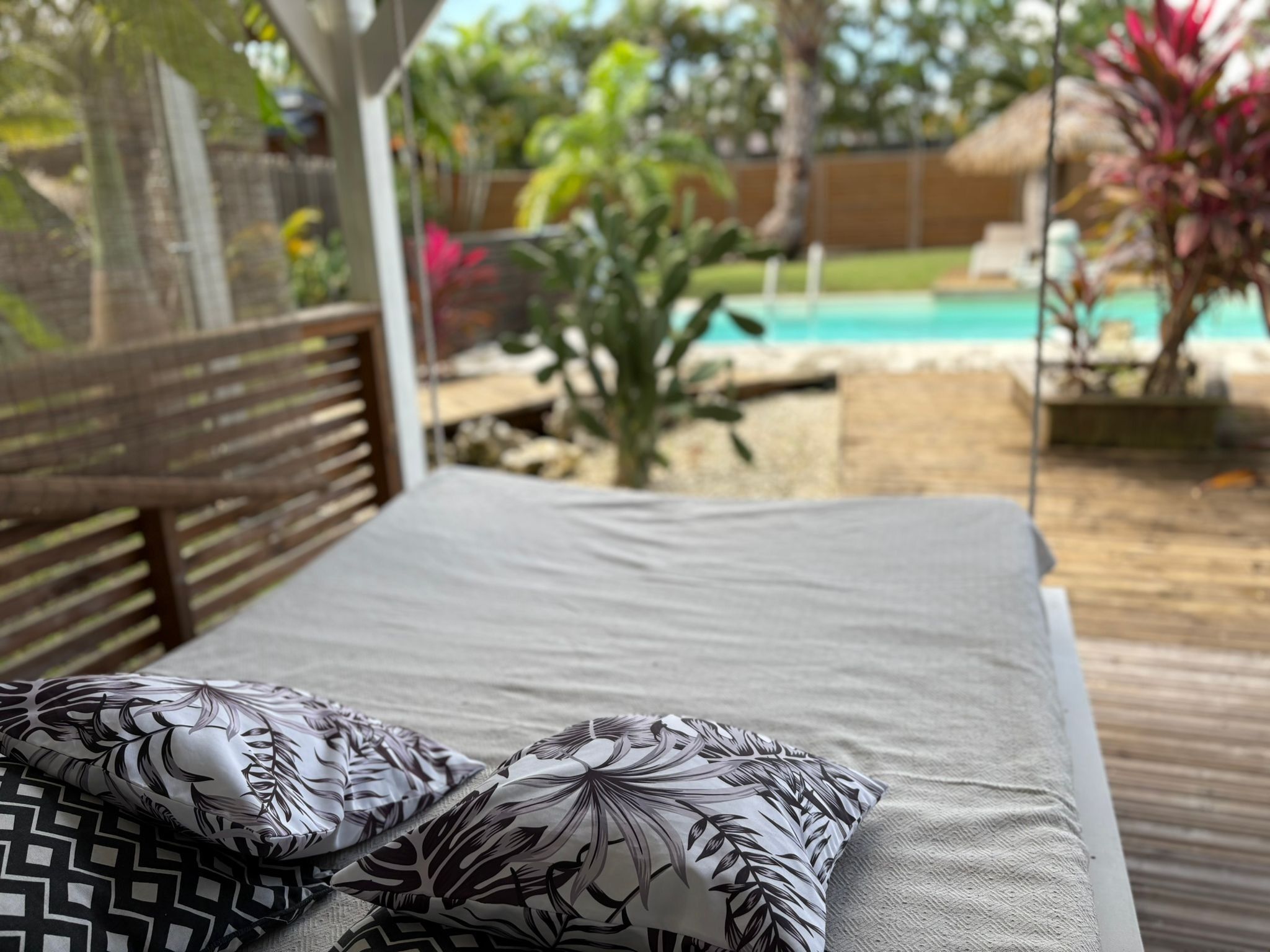 Ein Bett mit gemusterten Kissen, eine Holzterrasse und Blick auf Pool und Garten.