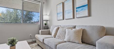 A modern living room with a sofa, a coffee table, and a potted plant. There are three framed artworks on the wall.