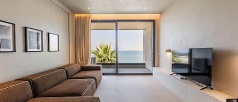 A modern living room with a sofa, a flat-screen TV, and a view of the sea through a sliding glass door.