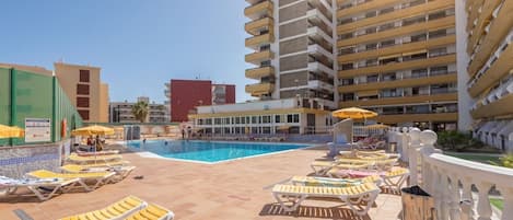 A pool area with sun loungers and umbrellas, surrounded by buildings.