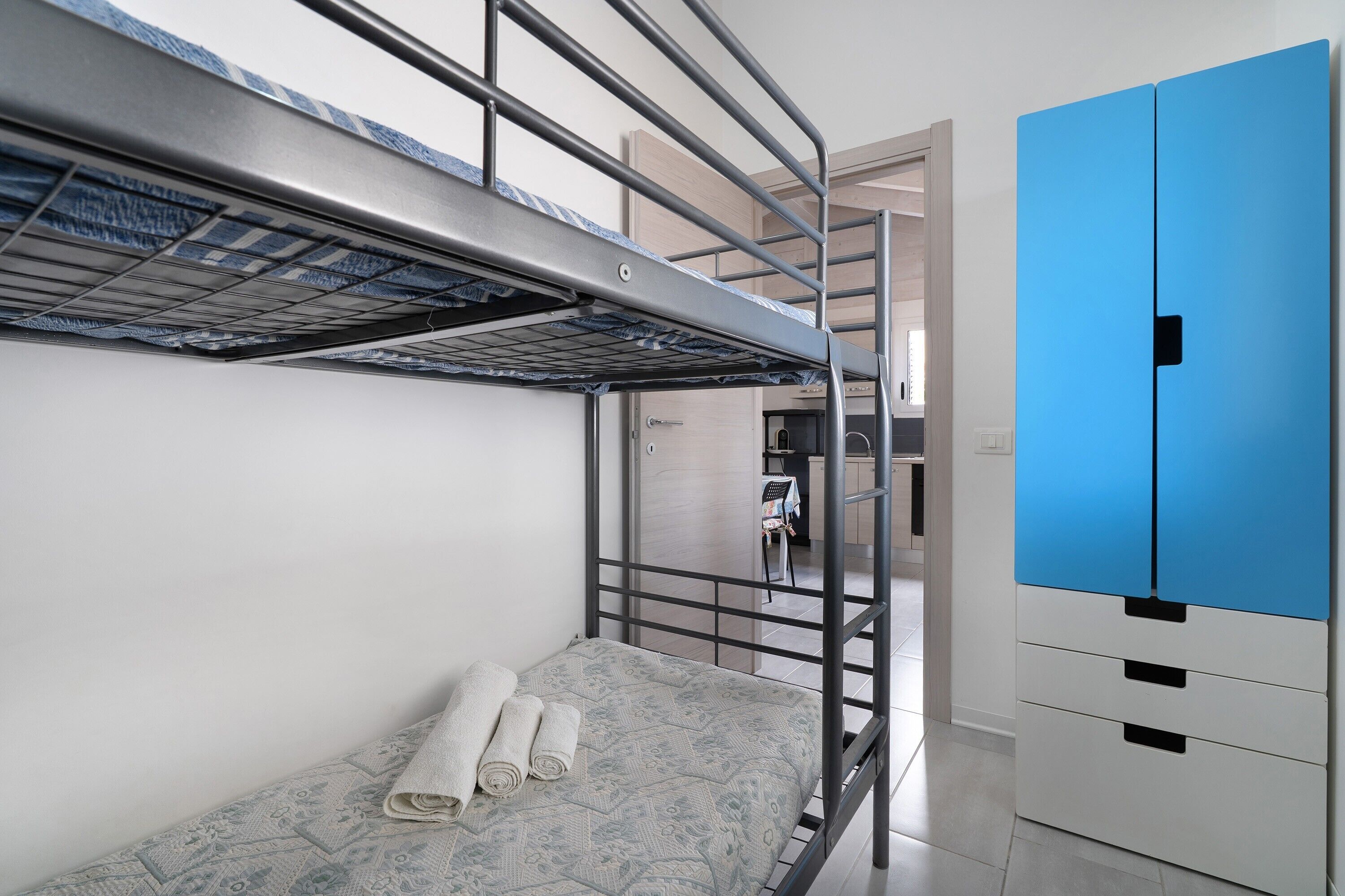 A bunk bed with a metal frame, a small table with a chair, and a blue wardrobe.