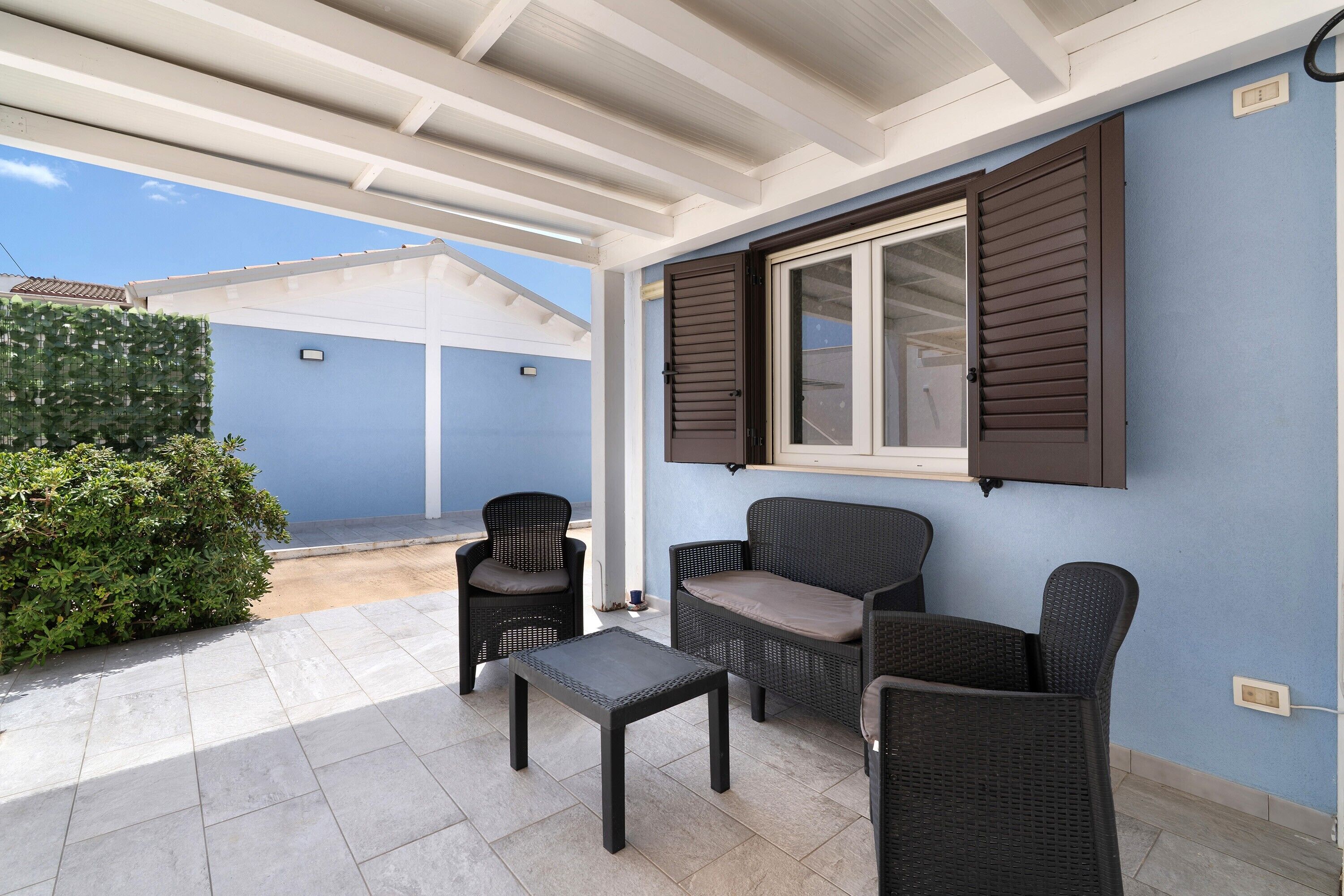 Ein überdachter Patio-Bereich mit Sitzgelegenheit, darunter ein Tisch und Stühle, sowie ein Fenster mit Rollläden.