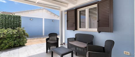 Un patio cubierto con un conjunto de mobiliario, incluyendo una mesa y sillas, y una ventana con contraventanas.