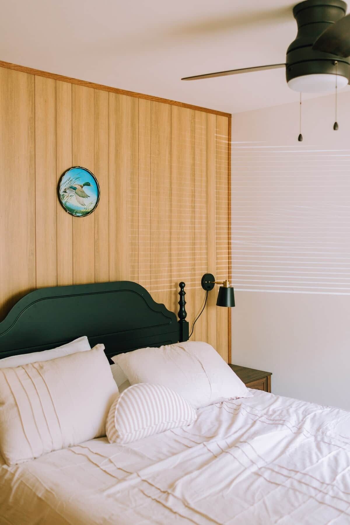 Ein Bett mit weißer Bettwäsche, einem grünen Kopfteil, einer Wanduhr, einem Deckenventilator und einer Nachttischlampe.