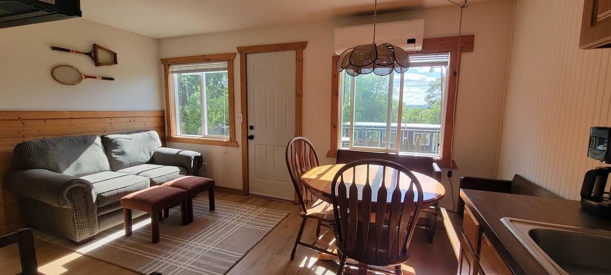 A cozy living room with a sofa, a dining table with chairs, a bench, and a kitchen area with a sink and a window.