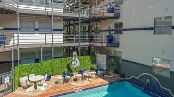 Piscina con sillas de descanso y sombrillas, rodeada por un edificio con varios balcones y vistas a un rascacielos.