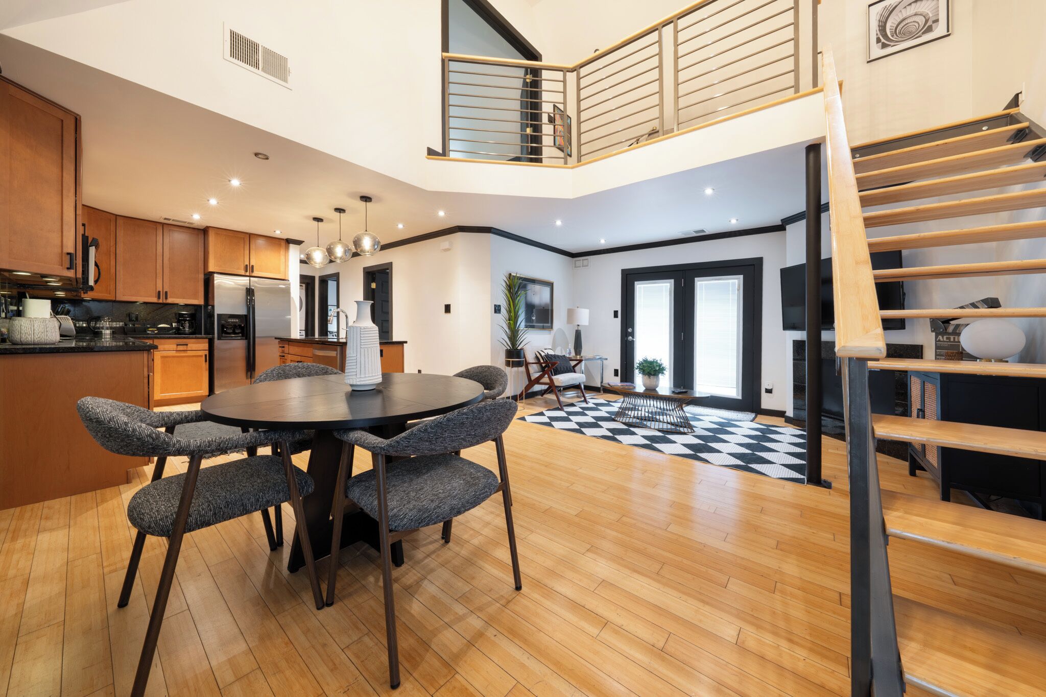 A modern kitchen with wooden cabinets, a dining area with a round table and chairs, and a staircase leading to a balcony.