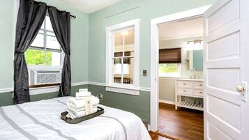 A hotel room with a bed, a window with curtains, an air conditioning unit, a bathroom with a sink and mirror, and a door leading to another room.
