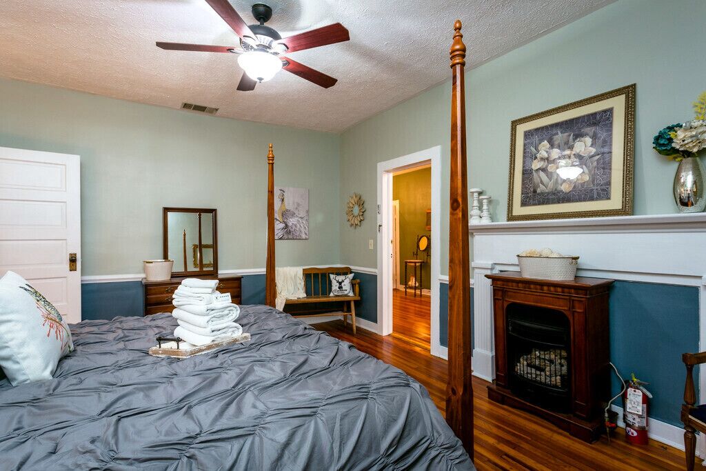A bedroom with a four-poster bed, a ceiling fan, a fireplace, and a door leading to another room.