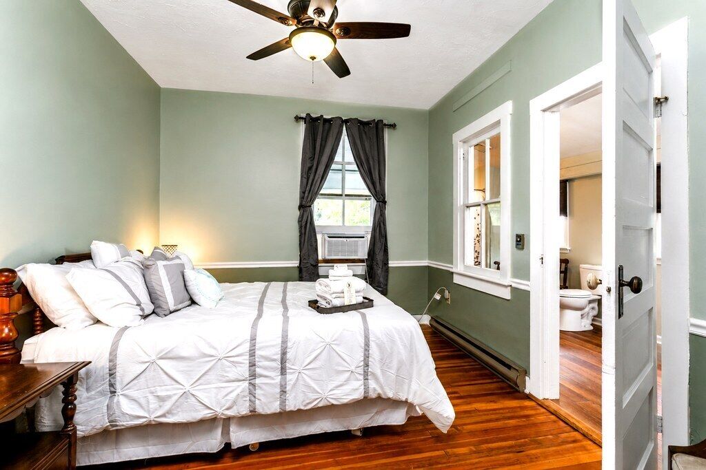 A bedroom with a bed, a ceiling fan, a window with curtains, a door, and a view of a bathroom.