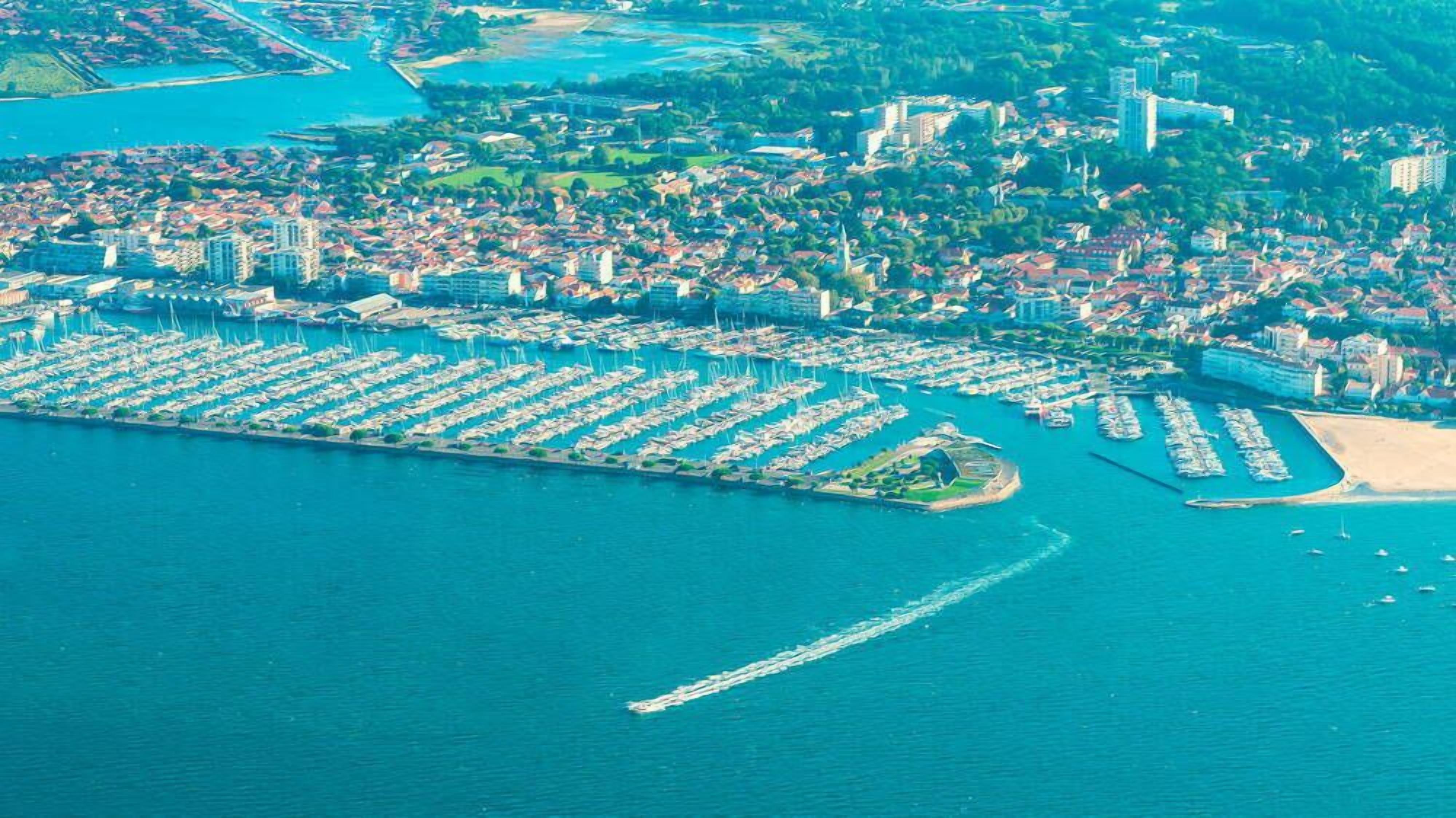 An aerial view of a coastal marina with numerous boats docked, surrounded by residential buildings and a sandy beach.