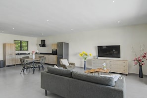 A modern living room with a grey sofa, a dining table with chairs, a kitchen area with a built-in oven, and a flat-screen TV mounted on the wall. - Felicidat Nobo, Mambo Beach (Willemstad)