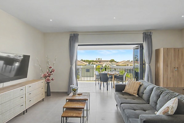 A living room with a flat-screen TV, a sofa, a dining table with chairs, and a balcony with a view of the neighborhood. - Felicidat Nobo, Mambo Beach (Willemstad)