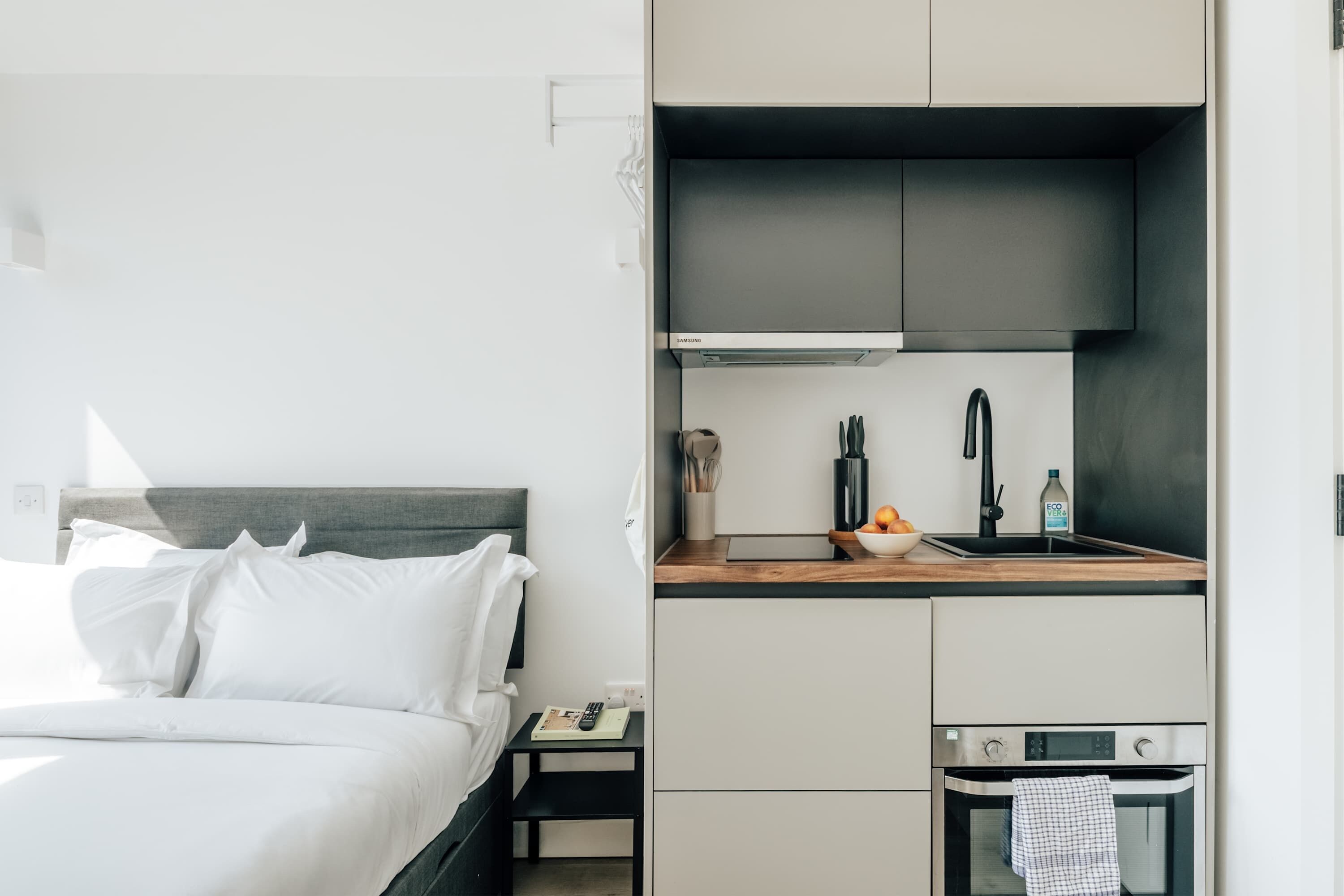 A compact hotel room with a bed, a small bedside table, a kitchenette with a stove, and a built-in oven.
