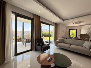 Ein geräumiges Wohnzimmer mit einer Couch, einem Sessel, einem Couchtisch und großen Fenstern mit Blick ins Freie.