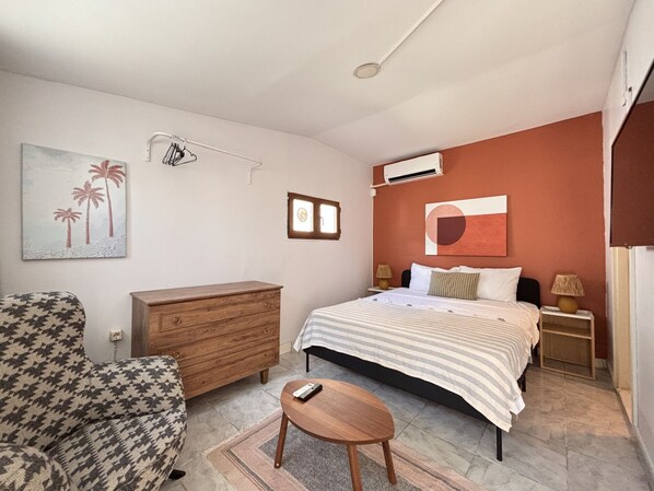 Ein Schlafzimmer mit einem Bett, einem hölzernen Schminktisch, einem Sessel, einer Klimaanlage und einem Bild an der Wand.