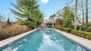 Una piscina rectangular con cubierta de madera, rodeada de árboles y una casa moderna al fondo.