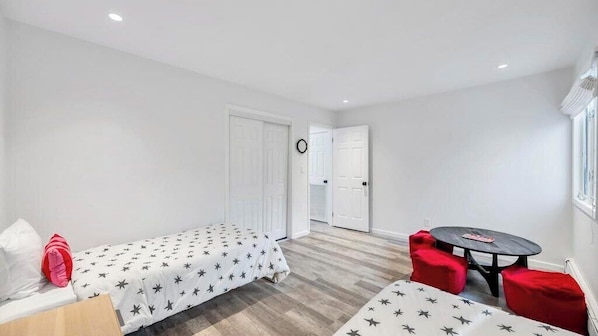 Une petite pièce bien éclairée, avec un lit, une table et deux chaises rouges. Une porte donne sur une autre pièce et une fenêtre se trouve sur le côté droit.