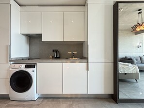 Eine moderne Küche mit weißen Schränken, einer eingebauten Waschmaschine und einem sichtbaren Wohnbereich mit Sofa und Bett.