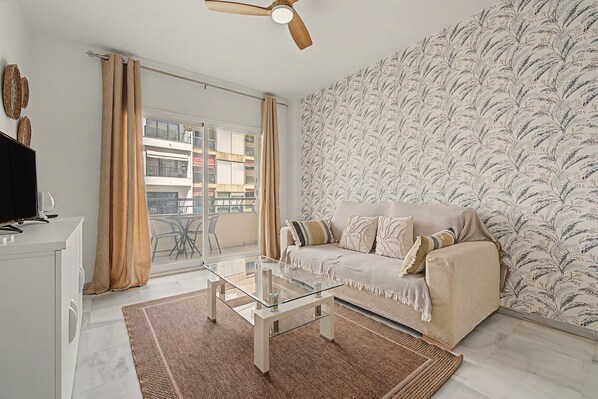 A living room with a sofa, a glass coffee table, a patterned wall, a ceiling fan, and a view of a balcony. - Apartment 'Gilarez' with Private Terrace, Balcony and Wi-Fi (Estepona)